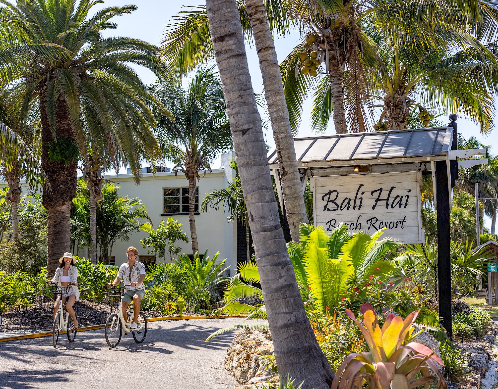 Two people ride bicycles near a tropical resort sign that reads 