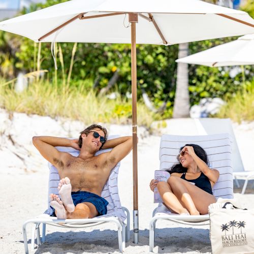 A couple relaxes on beach chairs under a white umbrella, with a beach bag nearby and vegetation in the background, ending the sentence.