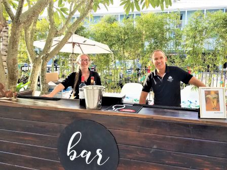 A wooden bar with a sign, two smiling bartenders, and some bar tools are set up outdoors in a garden-like area with trees and bamboo.