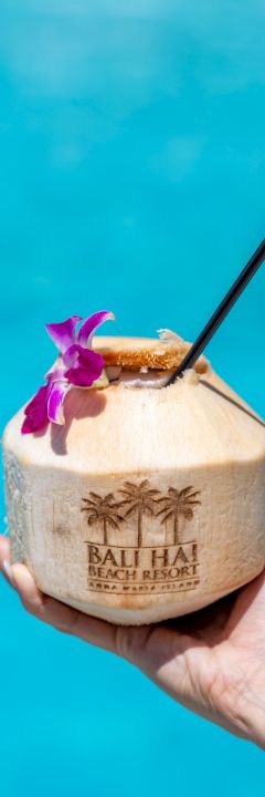 A hand holds a coconut drink adorned with a flower and straw against a blue water background. The coconut is labeled 