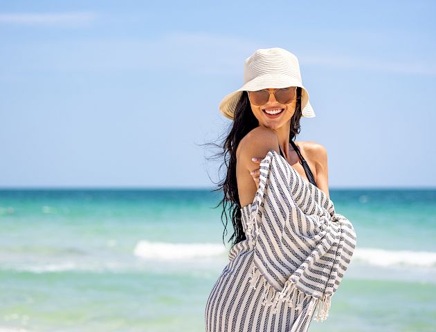 A smiling person wearing a striped dress and sun hat is posing on a beach with a blue ocean and sky in the background.